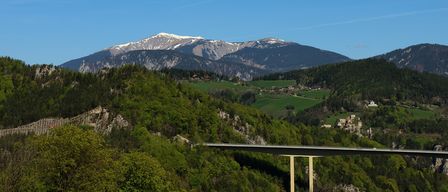 Eine Berglandschaft mit einer Brücke unter einem blauen Himmel mit schneebedeckten Gipfeln. Grüne Felder und Häuser säumen das Tal unten.