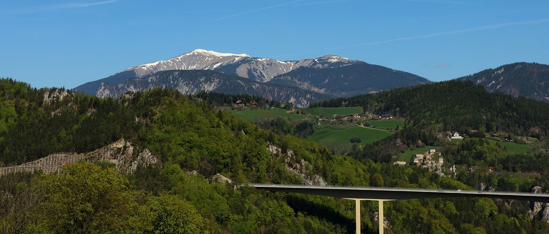 Eine Landschaft mit einer Brücke über einem Tal, mit schneebedeckten Bergen im Hintergrund und einem klaren blauen Himmel.