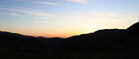 Die Sonne geht hinter einer Bergkette unter, mit einer Brücke darunter. Der Himmel ist mit flaumigen Wolken gefüllt.