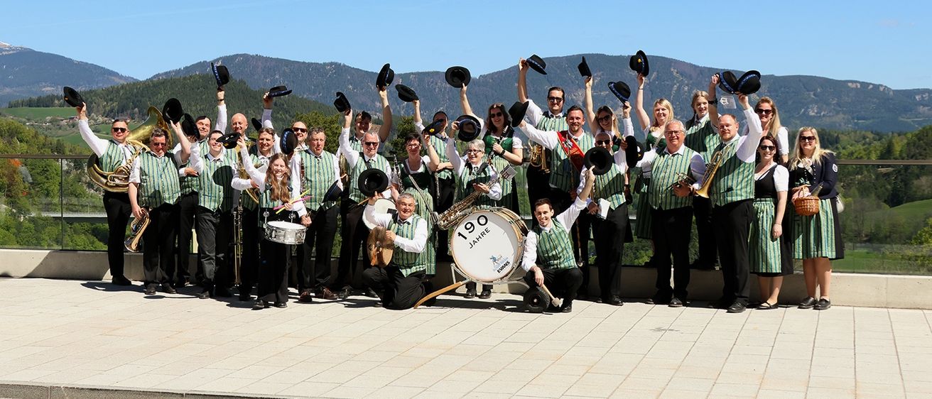 Ein Blasorchester posiert für ein Foto mit Bergen im Hintergrund. Die Mitglieder tragen grüne Westen, schwarze Hosen und Hüte. Sie halten Instrumente wie Trompeten, Posaunen und Trommeln.