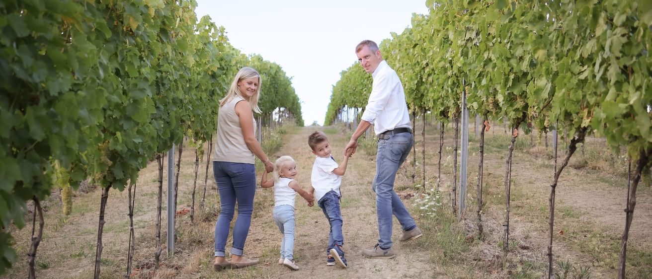 Eine vierköpfige Familie, bestehend aus zwei Eltern und zwei Kindern, geht einen Weinbergweg entlang. Die Eltern halten die Hände der Kinder.