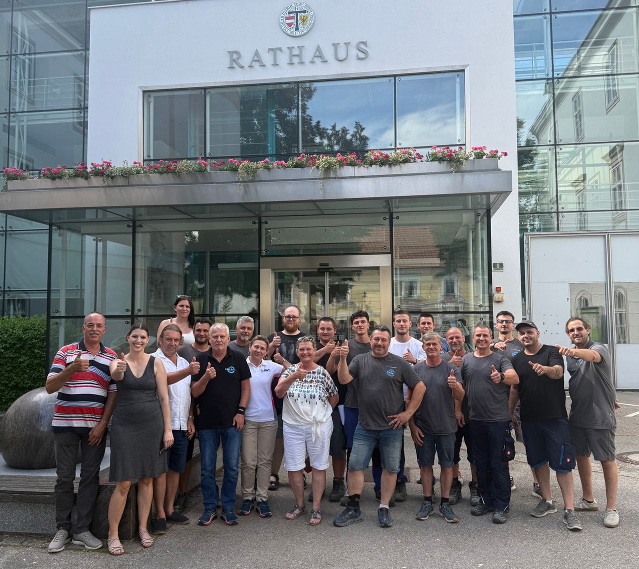 Eine Gruppe von Menschen, Männern und Frauen, steht vor einem Gebäude mit dem Wort Rathaus darauf. Sie alle zeigen Daumen hoch.