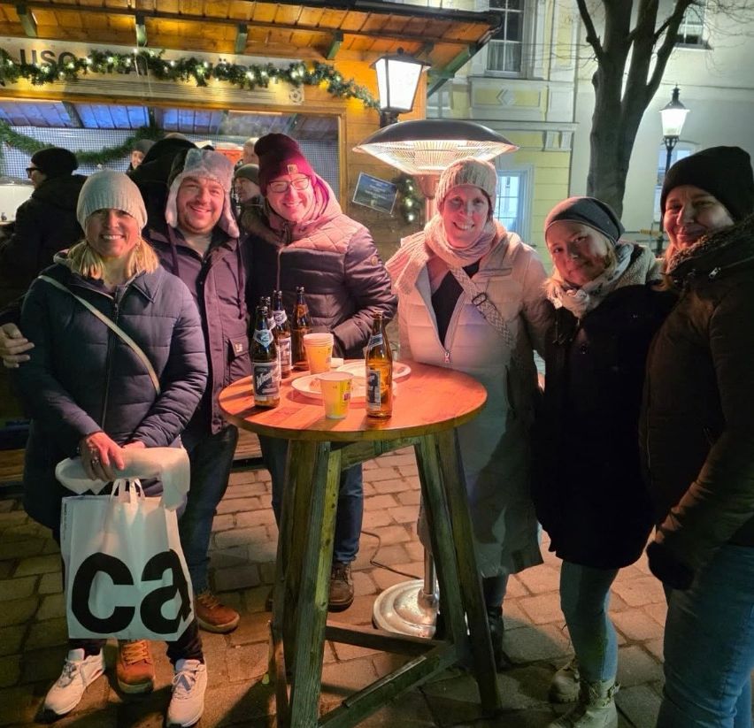 Sieben Personen stehen um einen Tisch herum vor einem Geschäft mit Getränken und einer Tasche. Sie tragen alle Winterkleidung. Hinter ihnen steht ein Gebäude mit einem Baum und einer Lampe.