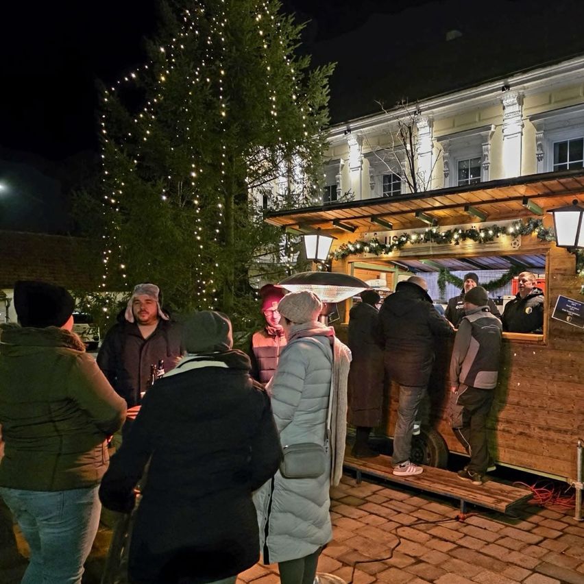 Bei Nacht stehen mehrere Personen vor einem Weihnachtsmarktstand, mit einem geschmückten Baum im Hintergrund.