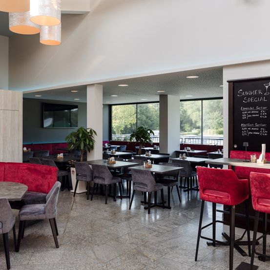 Ein Innenrestaurant mit Barbereich und mehreren Tischen. Es gibt rote gepolsterte Stühle und schwarze Barhocker. Eine Kreidetafel mit Speisekarte ist an der Wand angebracht.