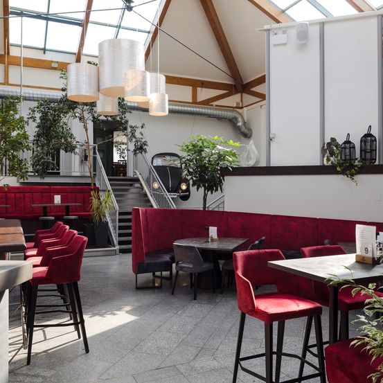 Ein Innenrestaurant mit hoher Decke und Glasdach. Es verfügt über rote Sitzgelegenheiten, runde Tische und hängende Lampen. Pflanzen und ein Oldtimer sind ebenfalls vorhanden.