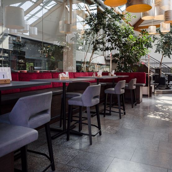 Ein Innenrestaurant mit hohen Decken, großen Glaswänden und modernem Design. Rote gepolsterte Bänke säumen die Wände, und graue Stühle sind um Tische herum angeordnet. Topfpflanzen und Lampen hängen von der Decke.