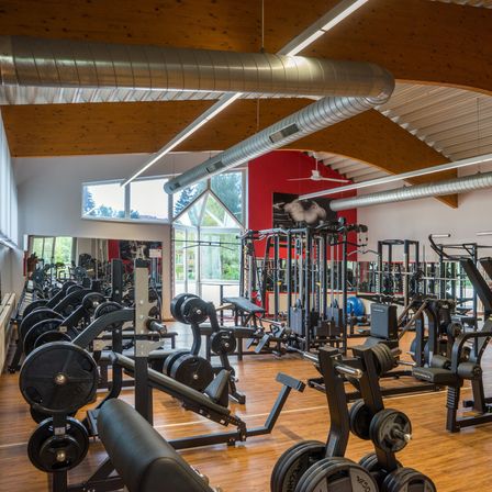 Ein gut ausgestattetes Fitnessstudio mit verschiedenen Trainingsgeräten und freien Gewichten, Holzböden und großen Fenstern, die natürliches Licht hereinlassen.