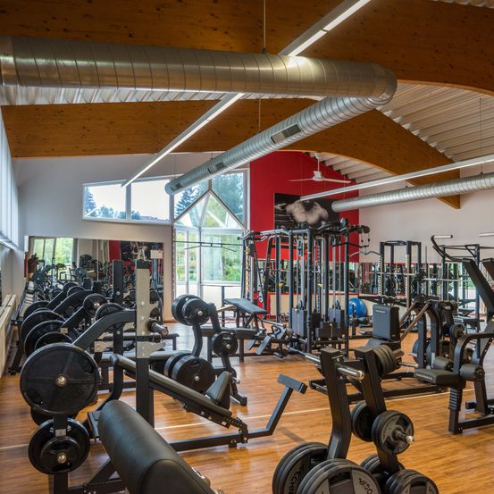 Ein gut ausgestattetes Fitnessstudio mit verschiedenen Trainingsgeräten und freien Gewichten, Holzböden und großen Fenstern, die natürliches Licht hereinlassen.