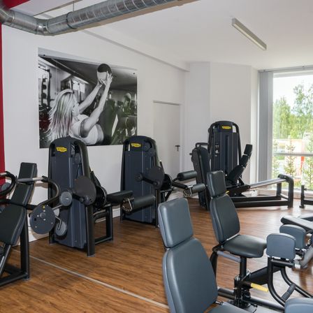 Ein Fitnessstudio mit einem großen Schwarz-Weiß-Foto an der Wand. Verschiedene Trainingsgeräte stehen auf Holzböden, und ein Glasfenster zeigt Bäume draußen.