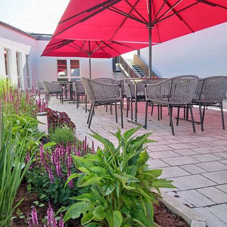Ein Patio mit Tischen und Stühlen, geschmückt mit roten Sonnenschirmen, ist von einem üppigen Garten mit lila Blumen und grünen Pflanzen umgeben.