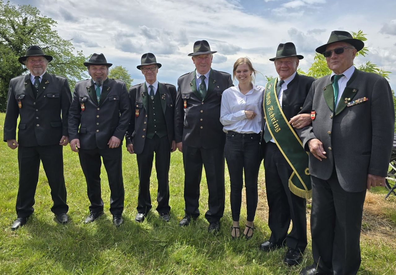 Eine Gruppe von Männern und einer Frau steht auf einem Feld, gekleidet in formelle Kleidung mit grünen Schärpen. Sie lächeln und posieren für ein Foto.