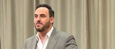 Ein Mann in einem Anzug steht auf der Bühne, gestikuliert und singt. Hinter ihm spielt ein anderer Mann Klavier. Sie stehen vor einem Vorhang und einem sitzenden Publikum.