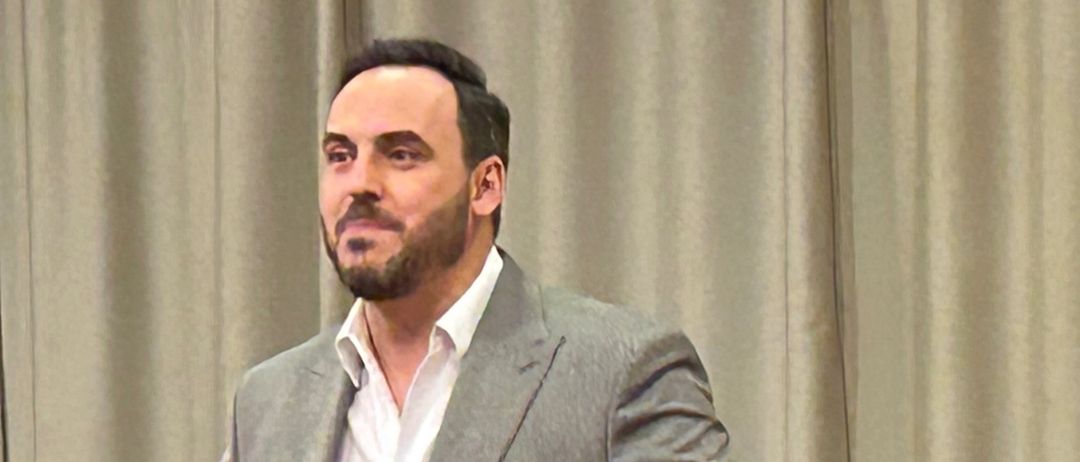 Ein Mann in einem Anzug steht auf der Bühne, gestikuliert und singt. Hinter ihm spielt ein anderer Mann Klavier. Sie stehen vor einem Vorhang und einem sitzenden Publikum.