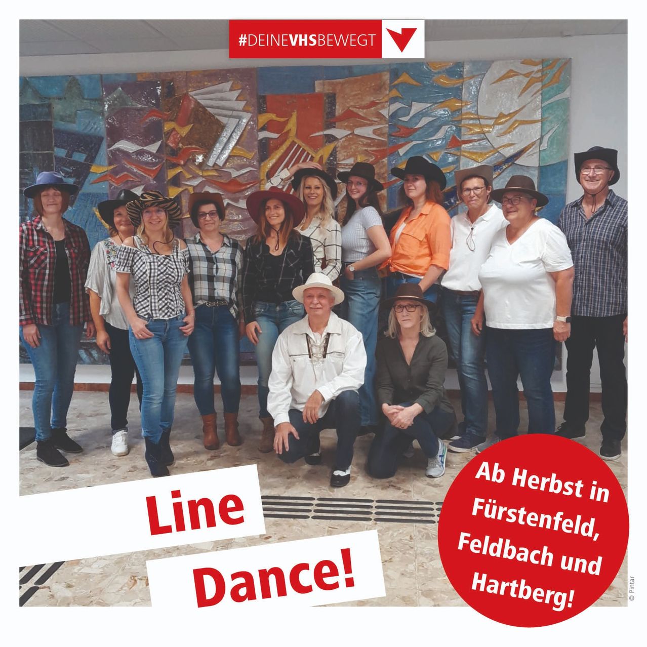 Gruppe von Menschen in Hüten posiert für ein Foto vor einem farbenfrohen Wandbild. Darunter stehen die Worte 'Line Dance!'