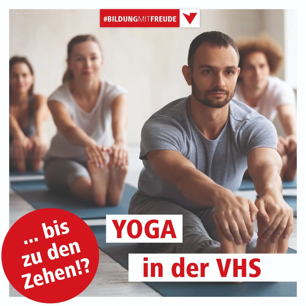 Eine Gruppe von Menschen praktiziert Yoga in einem Studio. Ein Mann vorne sitzt, während eine Frau neben ihm kniet. Die Worte Yoga in der VHS sind auf dem Teppich geschrieben.