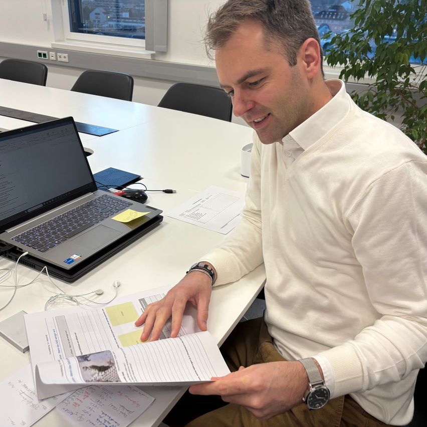 Ein Mann sitzt an einem Schreibtisch und liest lächelnd ein Dokument. Neben ihm steht ein Laptop mit Haftnotizen, und im Hintergrund zeigt ein Fenster eine Stadtlandschaft.