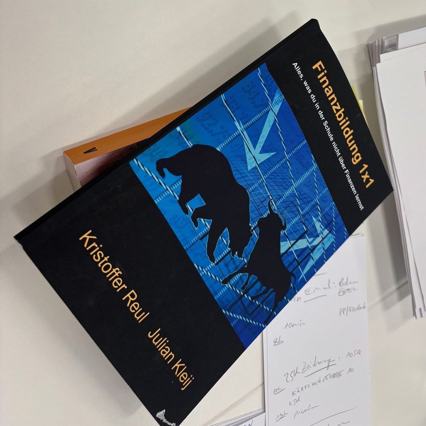 Das Buch mit dem Titel 'Finanzbildung x1' von Kristoffer Reul Julian Kleij liegt auf einer weißen Oberfläche. Es hat einen schwarzen Einband mit blauem Hintergrund und einer Grafik eines Bullen und eines Bären.