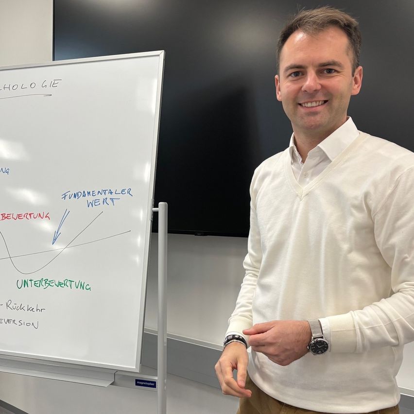 Ein Mann steht neben einer Tafel und lächelt. Die Tafel hat Diagramme und Text in verschiedenen Farben. Dahinter ist ein schwarzer Bildschirm zu sehen.