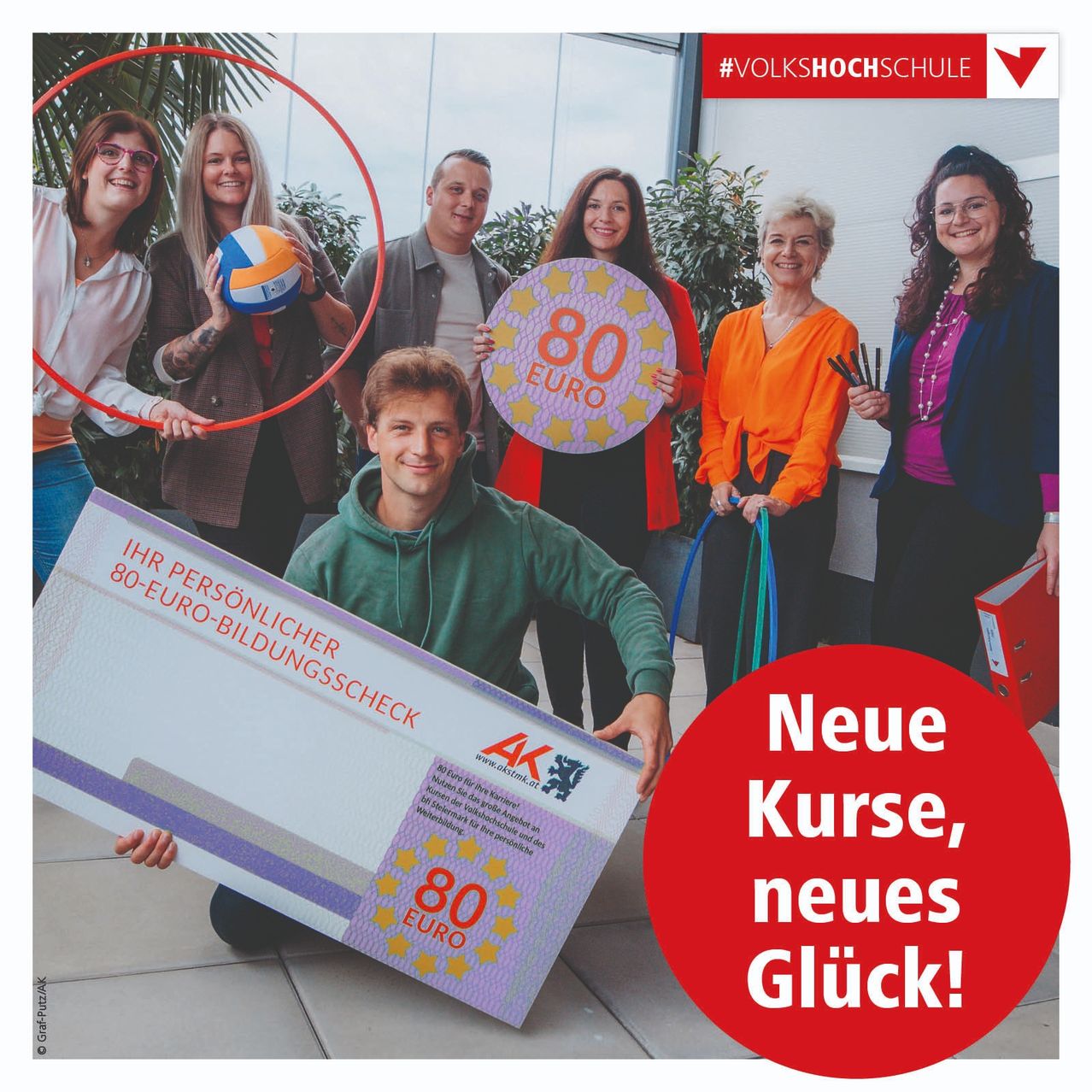 Eine Gruppe von Menschen steht um einen Mann, der ein Zertifikat und einen 80-Euro-Gutschein in der Hand hält. Der Text auf dem Gutschein lautet 'Neue Kurse, neues Gluck!'.