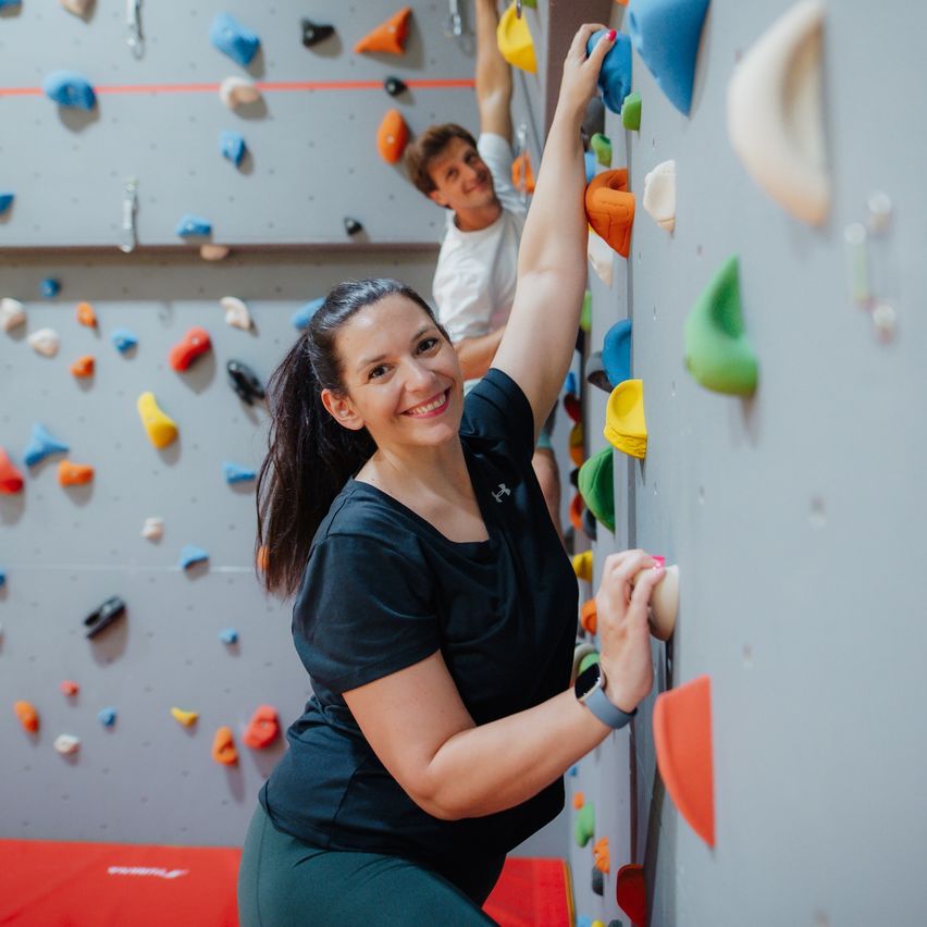 A woman and a boy are climbing on a wall with colorful grips. The woman is smiling and holding a grip.