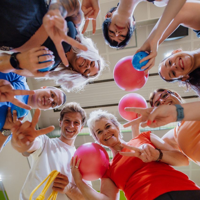 A group of people gathered in a circle, smiling and holding colorful balloons, appear to be participating in a fitness class or event.