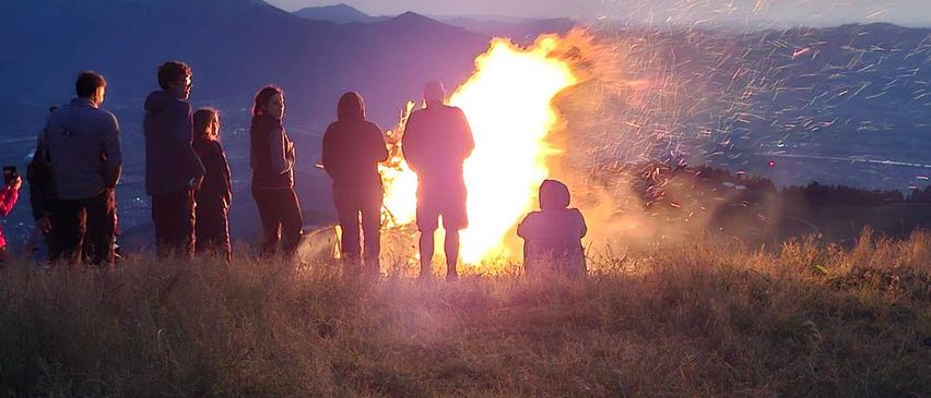 Eine Gruppe von Menschen steht um ein Lagerfeuer auf einem Hügel bei Sonnenuntergang. Die Flammen sind hell, und das Gebiet ist von Gras umgeben.