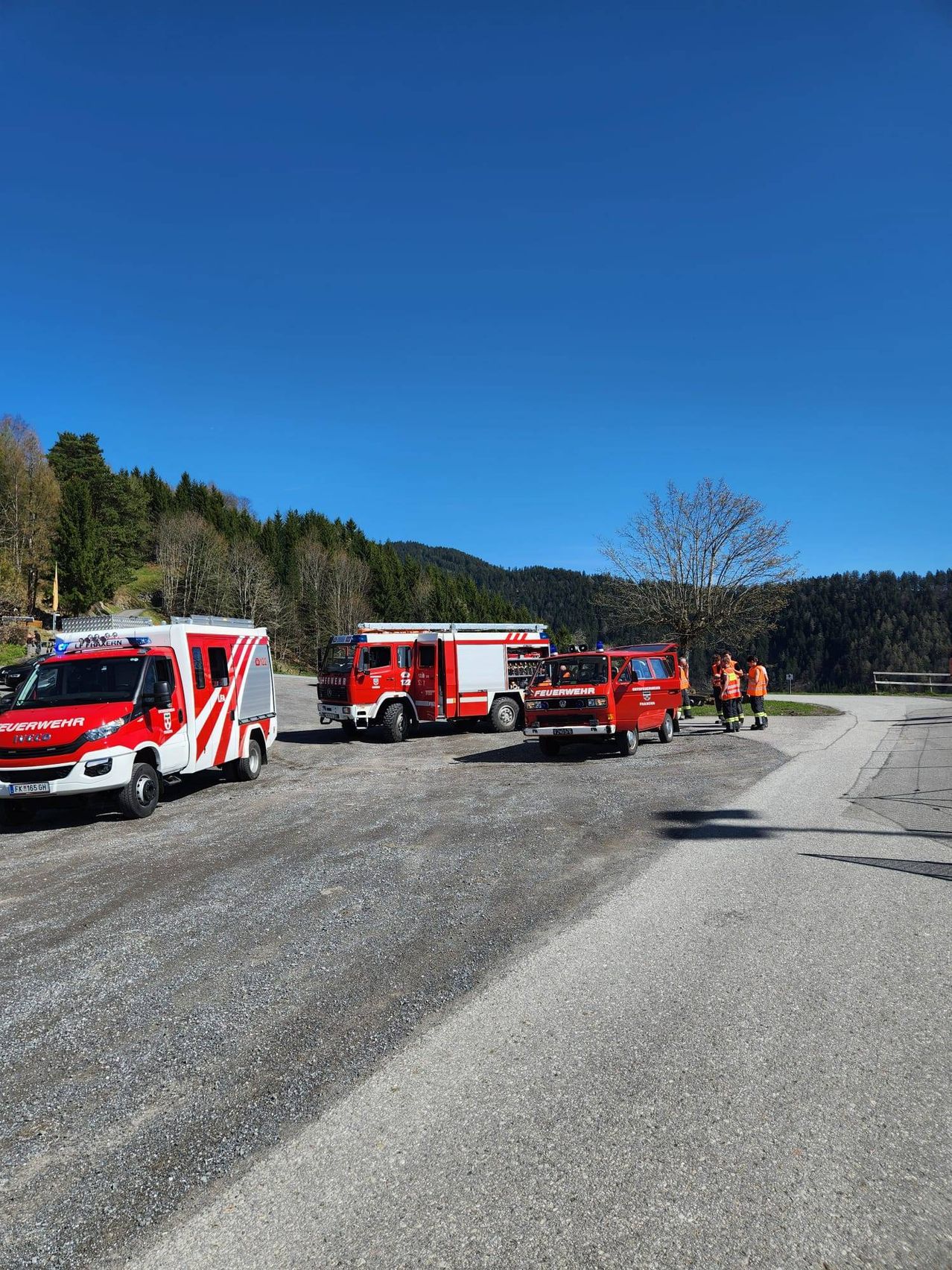 Three emergency vehicles are parked on a gravel road with mountains in the background. One vehicle has an open door.