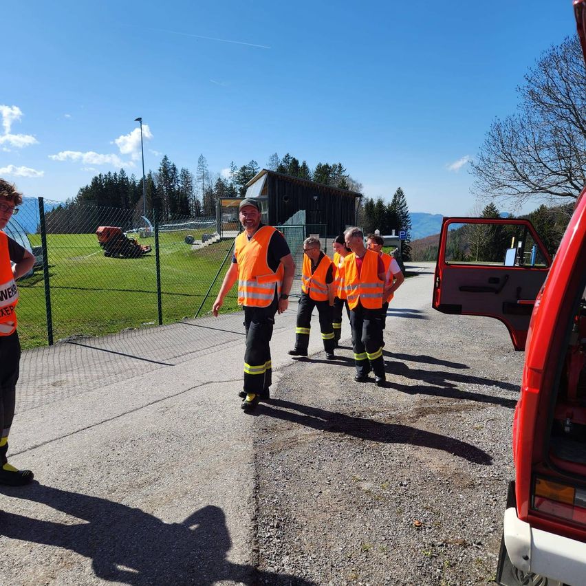 A group of people in orange vests stand near a vehicle with an open door on a gravel road. In the background is a fence, a field, and a building.