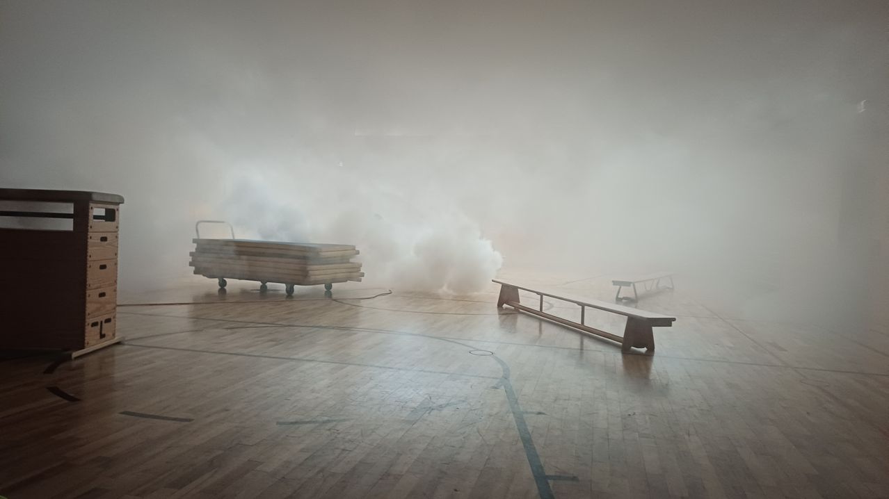 Ein Innenbereich mit Holzbänken, einem Stapel Holzbretter und dickem Nebel, der den Raum verdeckt.