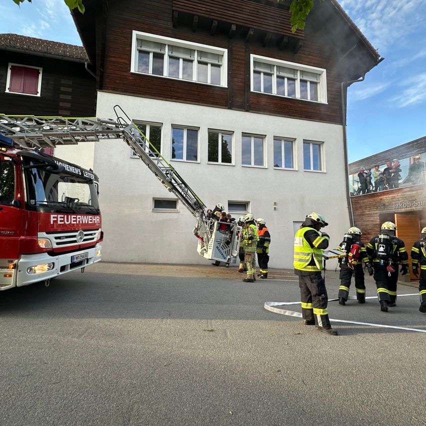 Ein Feuerwehrfahrzeug steht mit ausgefahrener Leiter vor einem Gebäude, aus dem Rauch aufsteigt. Feuerwehrleute sind in der Nähe versammelt.