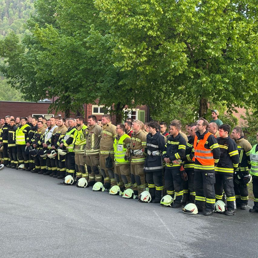 Eine Gruppe Feuerwehrleute in voller Uniform steht mit abgenommenen Helmen in einer Reihe, einige mit Westen, auf einer asphaltierten Straße. Dahinter stehen Bäume und ein Gebäude mit rotem Dach.