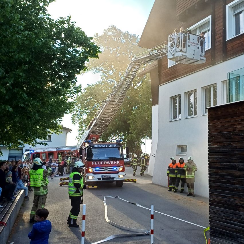Ein Feuerwehrauto ist auf der Straße geparkt, mit einer ausgefahrenen Leiter. Feuerwehrleute stehen um das Auto herum, und einige Leute beobachten vom Bürgersteig aus. Ein Gebäude befindet sich rechts von der Straße, und ein Baum steht links.