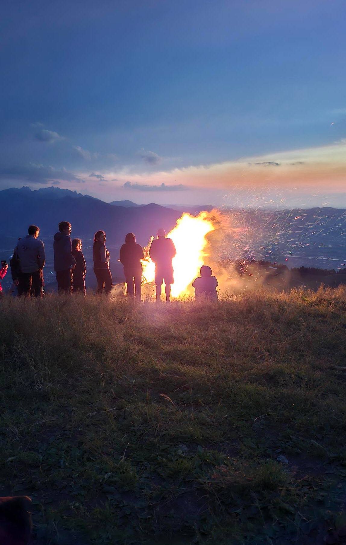 Eine Gruppe von Menschen steht um ein großes Lagerfeuer auf einem Hügel bei Sonnenuntergang, mit Bergen im Hintergrund.