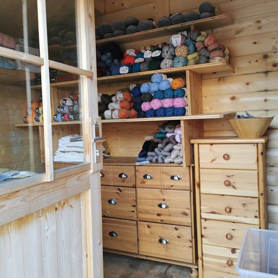 Ein kleiner Raum, gefüllt mit Holzregalen und -schubladen, die Garn in verschiedenen Farben und Stoffen enthalten. Eine Holztür ist leicht geöffnet, mit einer Schüssel rechts und einem Plastikbehälter links.