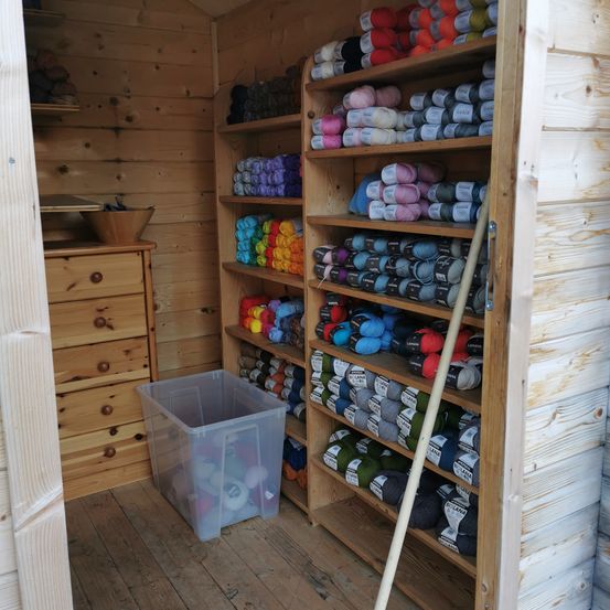 Ein hölzerner Raum mit Regalen voller verschiedener farbiger Garnrollen. Ein Plastikbehälter, gefüllt mit Garn, steht auf dem Boden. Ein Holzschrank befindet sich links, und ein Besen steht neben den Regalen.