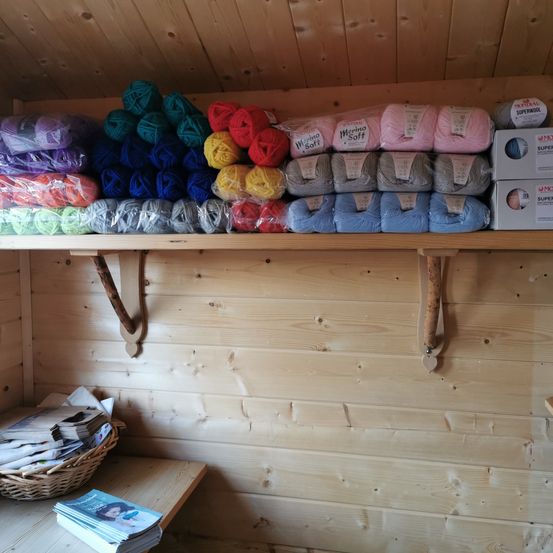 Ein Holzregal mit verschiedenen Garnen in unterschiedlichen Farben und Marken, einschließlich Merino Soft und Superwool, an einer Holzwand montiert. Darunter ein Korb mit Zeitschriften und ein Holztisch.