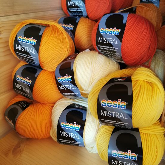 Eine Sammlung von Sesia Mistral Garnen in verschiedenen Farben, darunter Orange, Gelb und Weiß, ordentlich auf einer Holzoberfläche gestapelt.