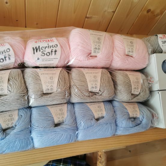 Verschiedene Farben von Merino Soft-Garnen in Plastiktüten auf einem Holzregal. Etiketten zeigen das Gewicht und die Zusammensetzung jedes Knäuels an.