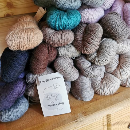 Ein Holzregal zeigt eine Vielzahl von Wollknäueln in verschiedenen Farben und Schattierungen. Ein weißes Schild mit dem Text 'Big Merino Hug' ist in der Mitte platziert.