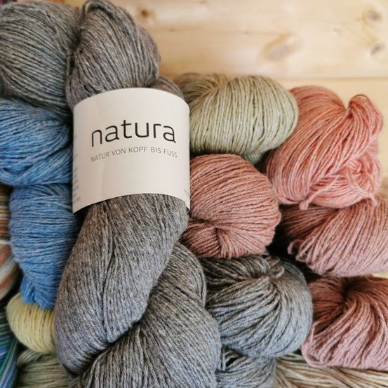 Ein Stapel Garne, darunter graue, blaue, rosa und grüne Knäuel, mit der Marke 'natura', präsentiert auf einem hölzernen Hintergrund.