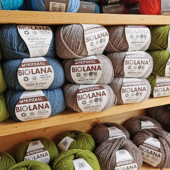 Ein Holzregal zeigt mehrere Wollknäuel, die als Bio Lana und Mondial gekennzeichnet sind und alle in Italien hergestellt werden. Die Wolle ist in verschiedenen Farben und Größen erhältlich.