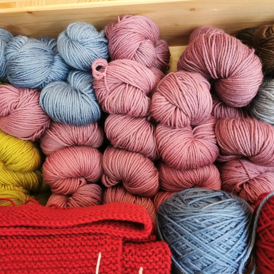 Ein Holzkasten gefüllt mit verschiedenen farbigen Garnen, darunter Blau, Pink, Gelb und Braun. Ein rotes gestricktes Objekt liegt neben den Garnen.