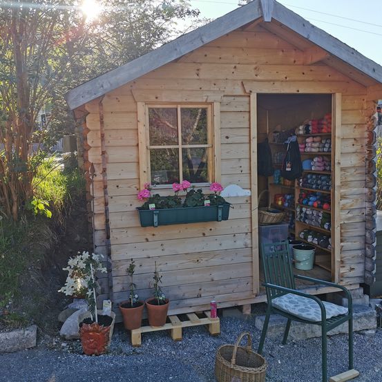 Eine kleine Holzhütte mit Fenster, Tür und Pflanzen draußen. Drinnen steht ein grüner Stuhl und ein Korb.