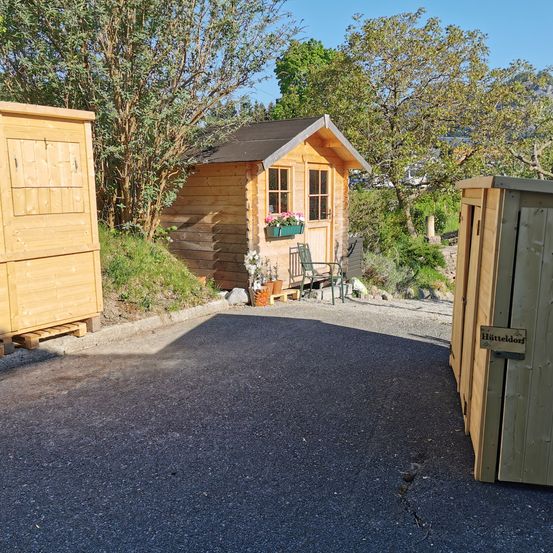 Ein kleines Holzhaus mit Blumenkasten, einem Stuhl und einer Bank an der Seite. Es gibt zwei Holzlagerboxen links und rechts. Bäume und Pflanzen sind im Hintergrund.