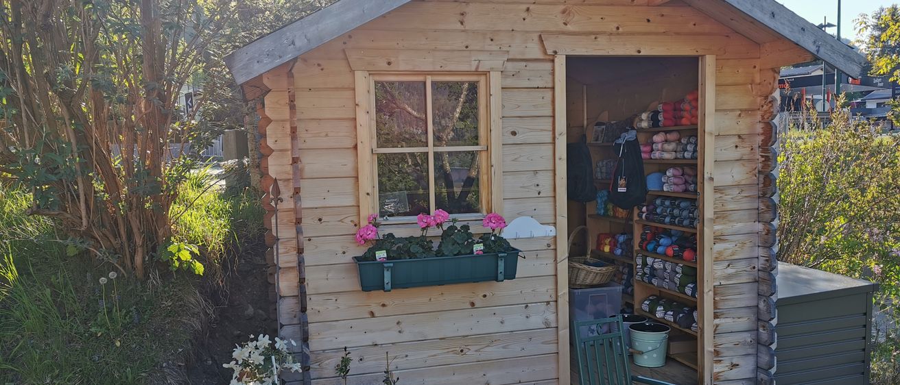 Eine kleine Holzhütte mit Fenster und offener Tür, die einen Blumenkasten, Topfpflanzen und einen Stuhl zeigt. Das Innere ist mit verschiedenen Gegenständen gefüllt, einschließlich eines Korbes, Eimern und farbenfrohen Artikeln.
