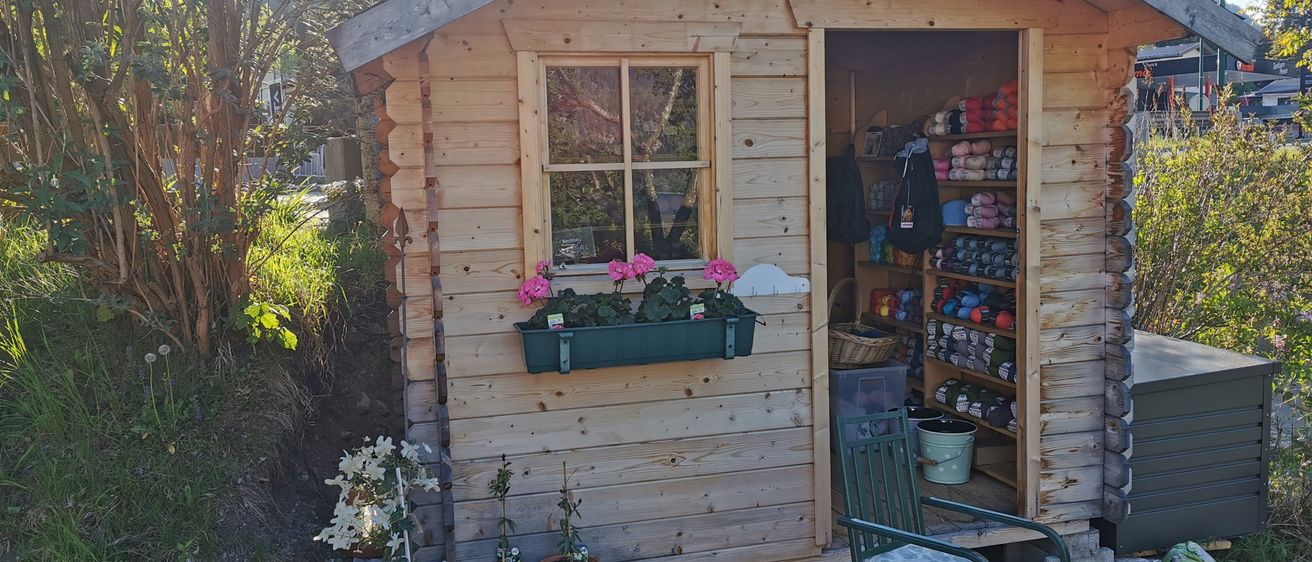 A small wooden cabin with a window and open door, showcasing a flower box, potted plants, and a chair. The interior is filled with various items, including a basket, buckets, and colorful items.