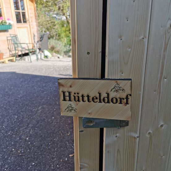 A wooden sign with the word Hutteldorf attached to a wooden wall. The wall is on a street with a parked car.