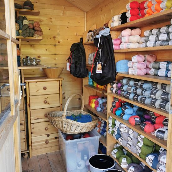A wooden room filled with shelves stocked with various yarn colors. A basket and plastic container are placed on the floor. A wooden cabinet and a bowl are on the left.
