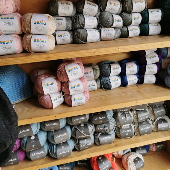 A wooden shelf displays various yarns in different colors and brands. 'Sesia' and 'Lamana' are prominently featured. The yarns are organized in rows on the shelf.